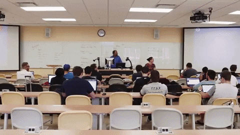 A classroom setting with an instructor presenting to his students listening while on their desks.