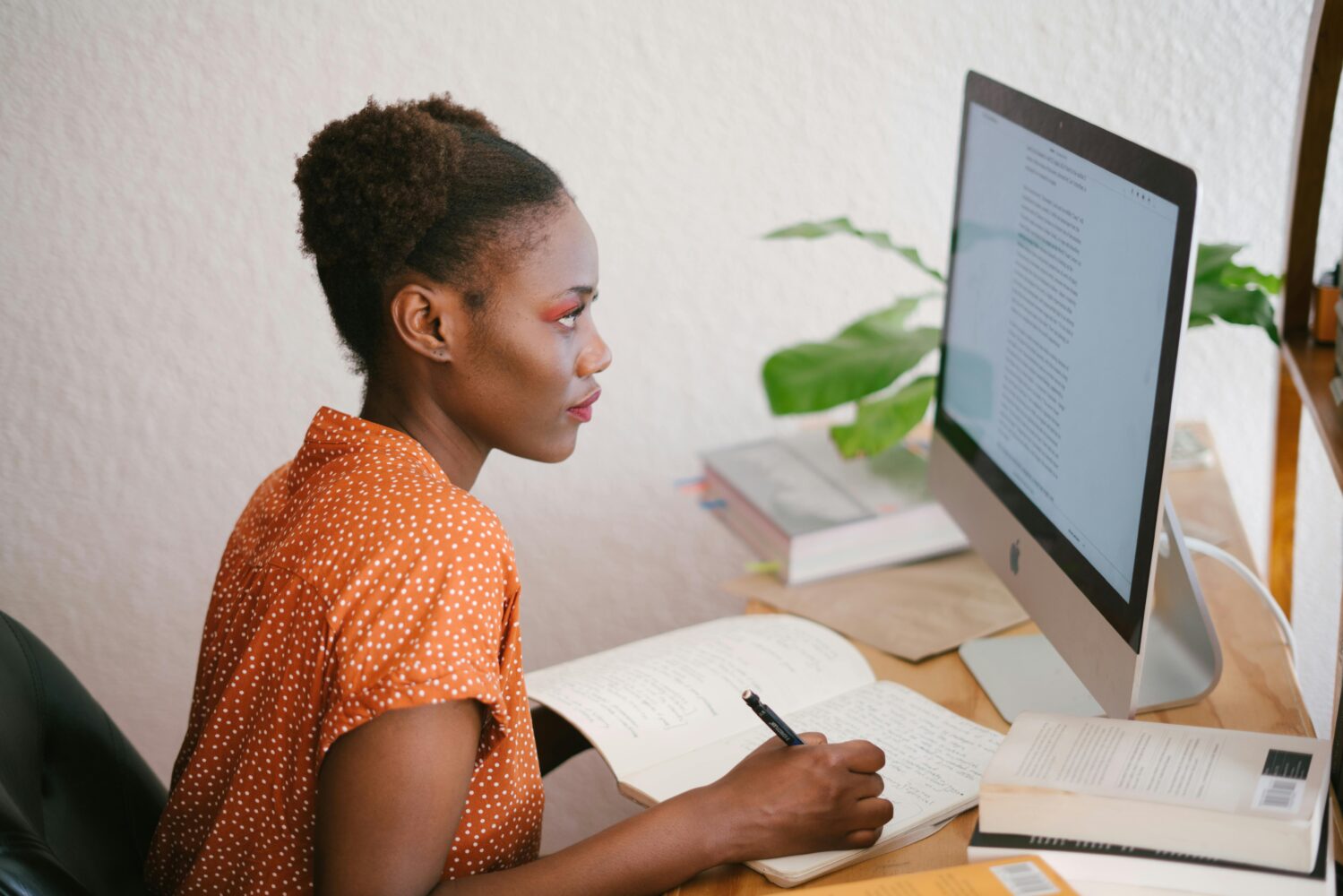 Woman in oragne top writing on desk in front of a computer screen