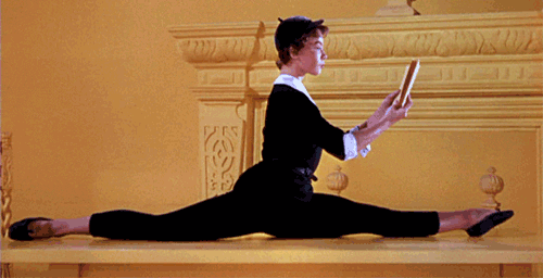 A woman in black suit reading a book while doing splits on the floor.