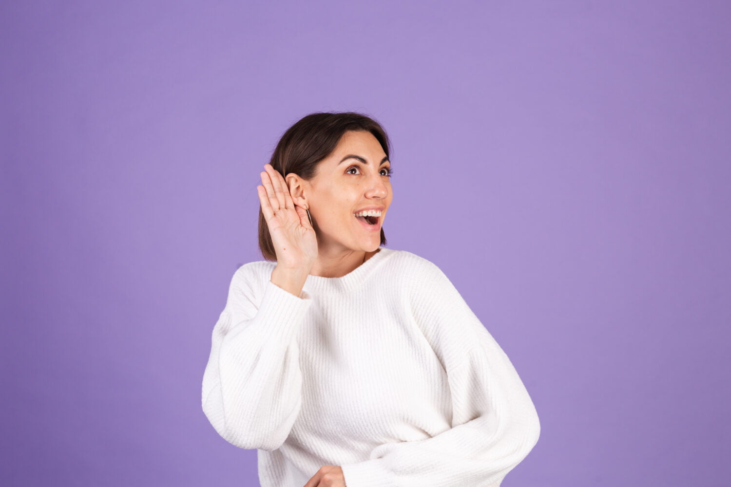 Young brunette in white casual sweater isolated on purple background curious try to hear what you saying with hand by ear copy space