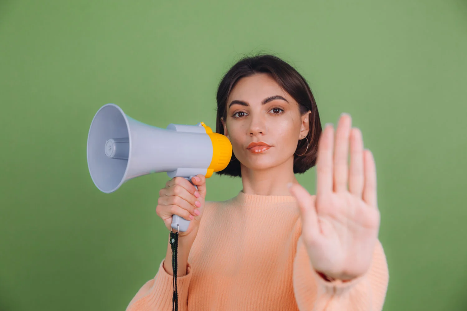 Speaking in Public: How to Use the Power of the Pause for Memorable Talks 4 young woman casual peach sweater isolated green olive color wall unhappy serious with megaphone doing stop sing with palm hand 1 Speaking in Public: How to Use the Power of the Pause for Memorable Talks